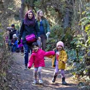 2 preschoolers walk together at Tree Time with their mothers following in the background.